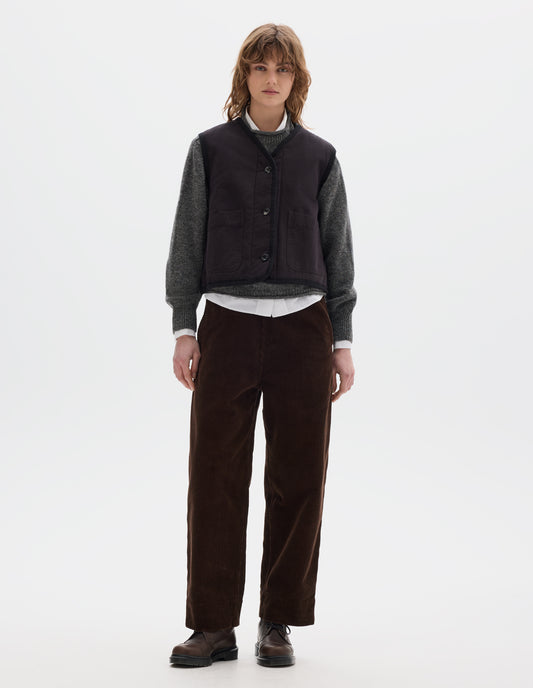 Full body image, front view of model wearing a black cotton sleeveless lightly padded liner with two front patch pockets and herringbone binding along all edges and seams. Styled with a grey crew wool sweater, worn over a white shirt, with brown corduroy trousers and brown leather shoes.