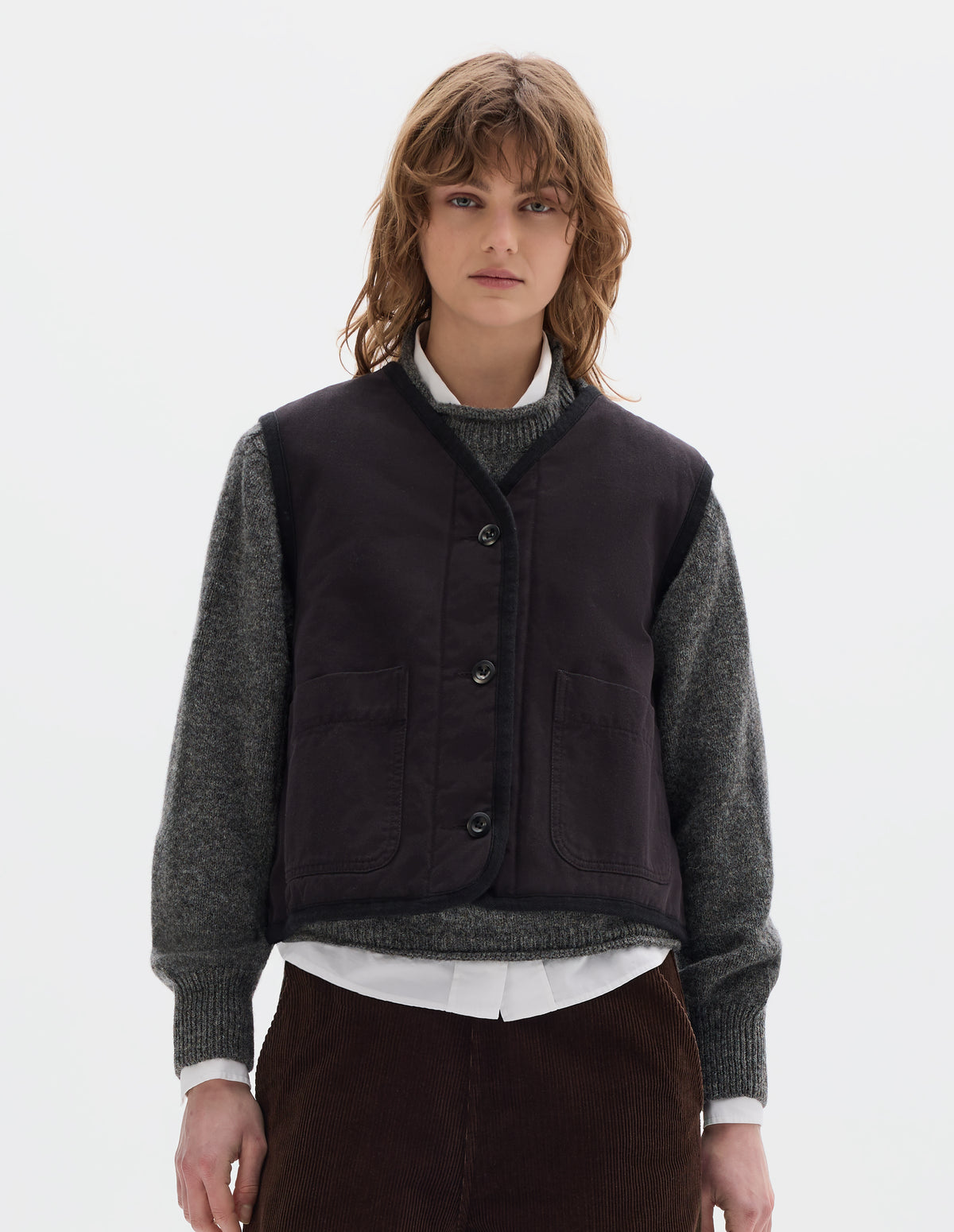 Cropped body image, front view of model wearing a black cotton sleeveless lightly padded liner with two front patch pockets and herringbone binding along all edges and seams. Styled with a grey crew wool sweater, worn over a white shirt, with brown corduroy trousers.
