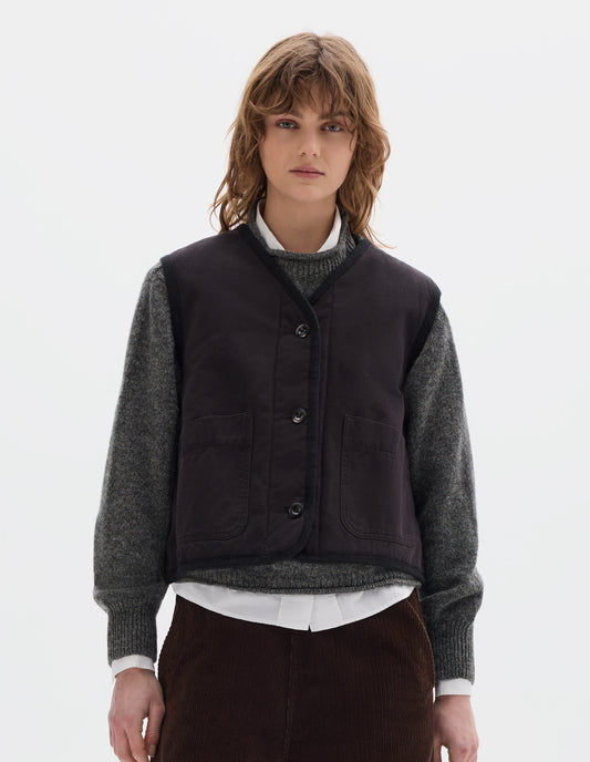 Cropped body image, front view of model wearing a black cotton sleeveless lightly padded liner with two front patch pockets and herringbone binding along all edges and seams. Styled with a grey crew wool sweater, worn over a white shirt, with brown corduroy trousers.