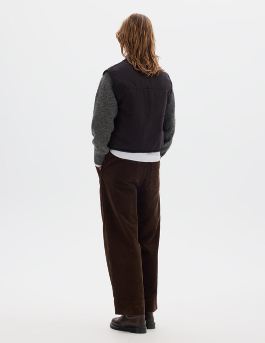 Full body image, back view of model wearing a black cotton sleeveless lightly padded liner with herringbone binding along all edges and seams. Styled with a grey crew wool sweater, worn over a white shirt, with brown corduroy trousers and brown leather shoes.