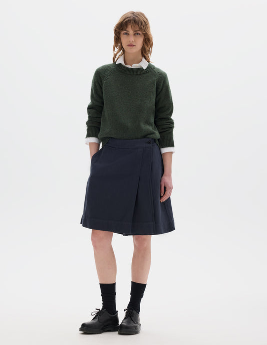 Full body image, front view of model wearing a dark green crew neck jumper over a white collared shirt, styled with a dark navy wrap skirt with concealed shorts with dark button fastening. Model is wearing black leather shoes with black over the ankle socks.