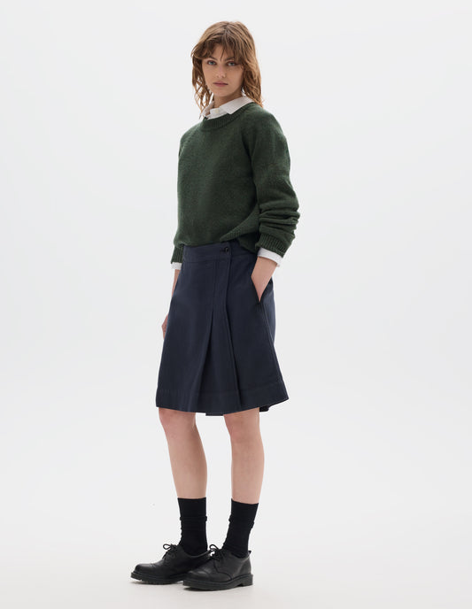 Full body image, side view of model wearing a dark green crew neck jumper over a white collared shirt, styled with a dark navy wrap skirt with concealed shorts with dark button fastening and side slant pocket. Model is wearing black leather shoes with black over the ankle socks.