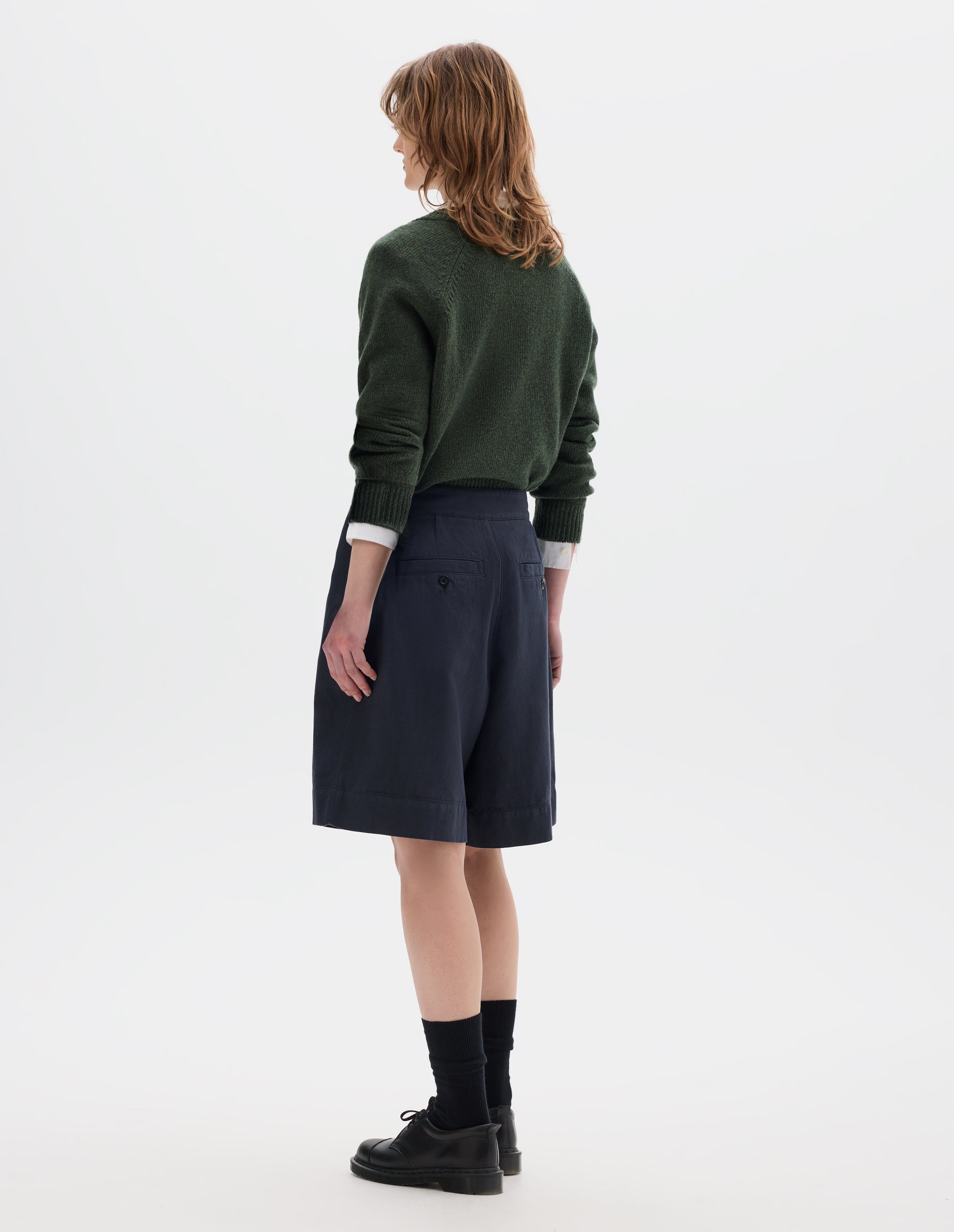 Full body image, back view of model wearing a dark green crew neck jumper over a white shirt, styled with a dark navy wrap skirt with concelaed shorts with two button-fastening welt pockets. Model is wearing black leather shoes with black over the ankle socks.