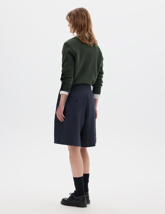 Full body image, back view of model wearing a dark green crew neck jumper over a white shirt, styled with a dark navy wrap skirt with concelaed shorts with two button-fastening welt pockets. Model is wearing black leather shoes with black over the ankle socks.