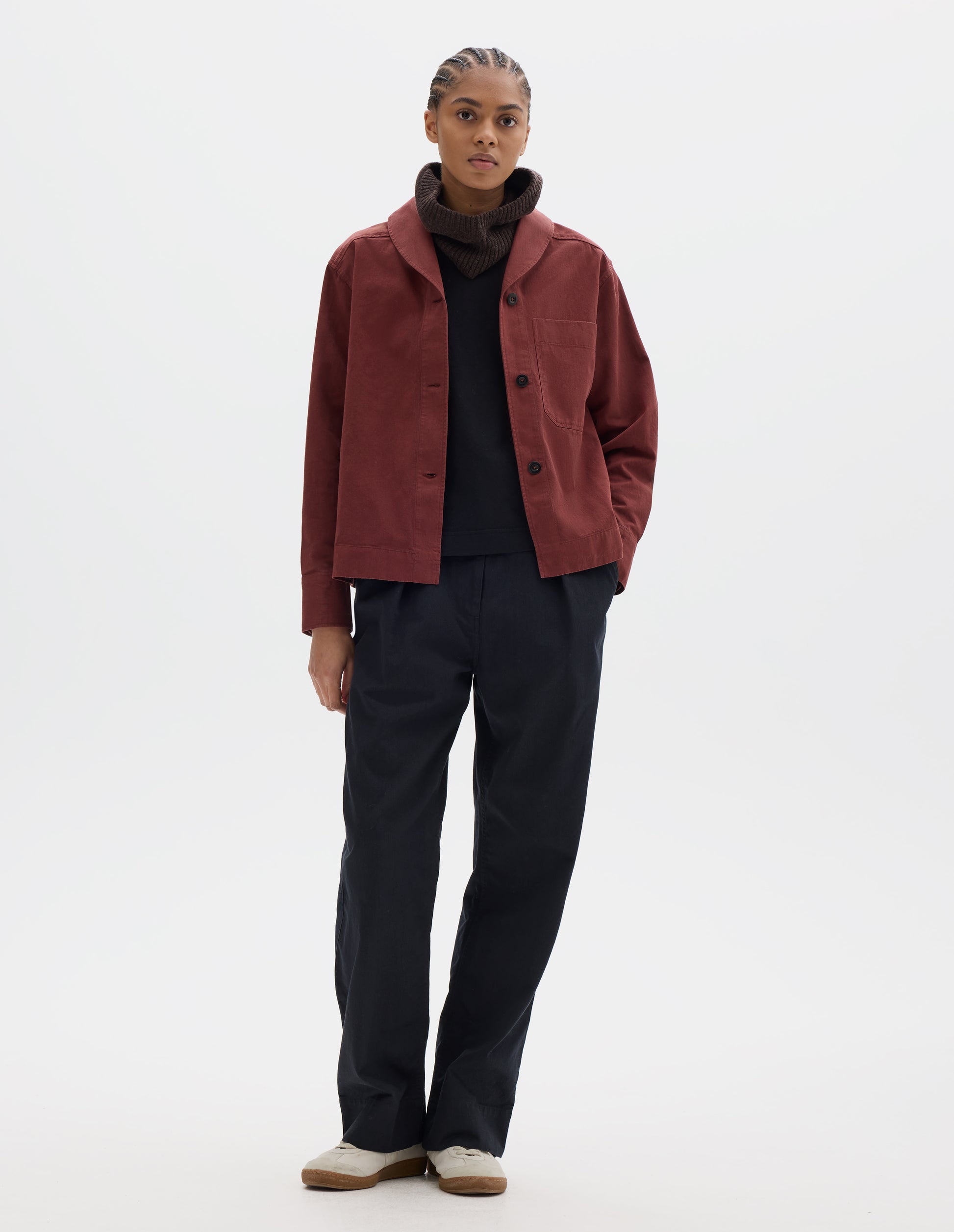 Full body image, front view of model wearing a reddish cotton shawl collar overshirt with a front button placket, dark buttons, a forward-positioned shoulder seam and a front patch pocket. Styled with a brown wool hood, black t shirt and pleated trousers, off white suede trainers.