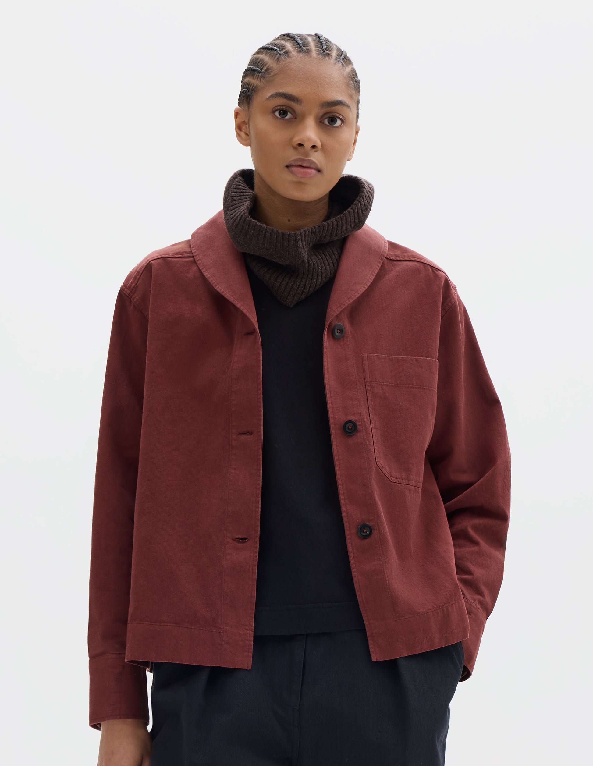 Cropped body image, front view of model wearing a reddish cotton shawl collar overshirt with a front button placket, dark buttons, a forward-positioned shoulder seam and a front patch pocket. Styled with a brown wool hood, black t shirt and pleated trousers.