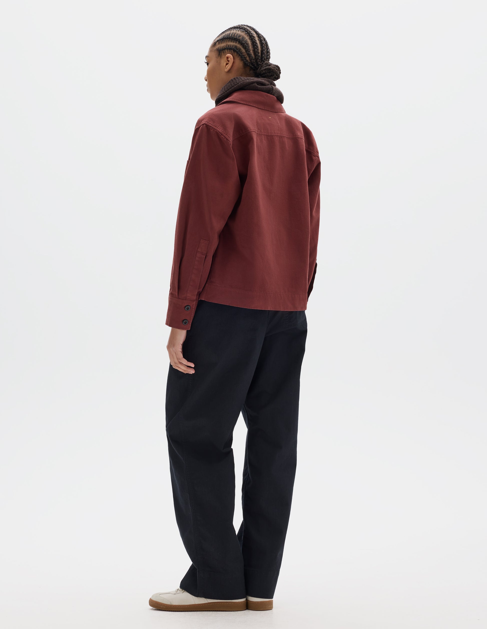 Full body image, back view of model wearing a reddish cotton shawl collar overshirt with a forward-positioned shoulder seam, back yoke detailing and cuffs with double button fastening. Styled with a brown wool hood, black pleated trousers and off white suede trainers.