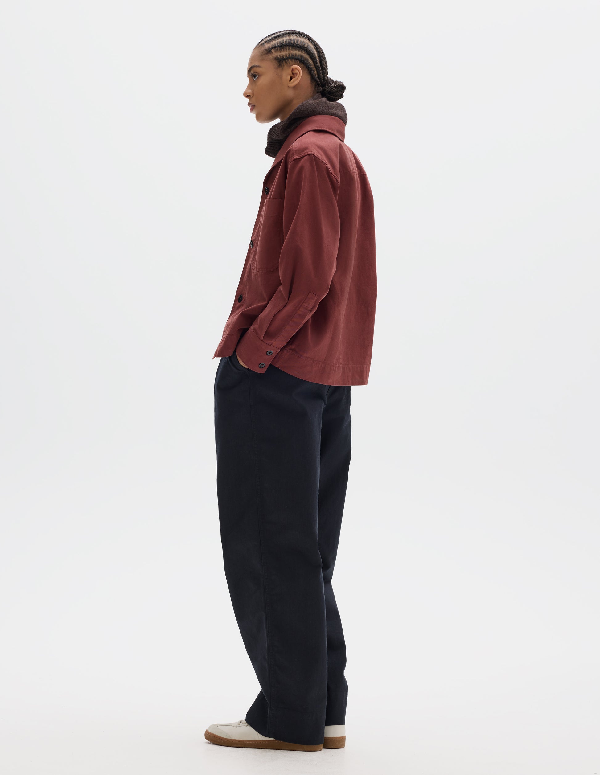 Full body image, front side view of model wearing a reddish cotton shawl collar overshirt with a front button placket, a forward-positioned shoulder seam and shirt cuffs with double button fastening. Styled with a brown wool hood, pleated trousers, off white suede trainers.