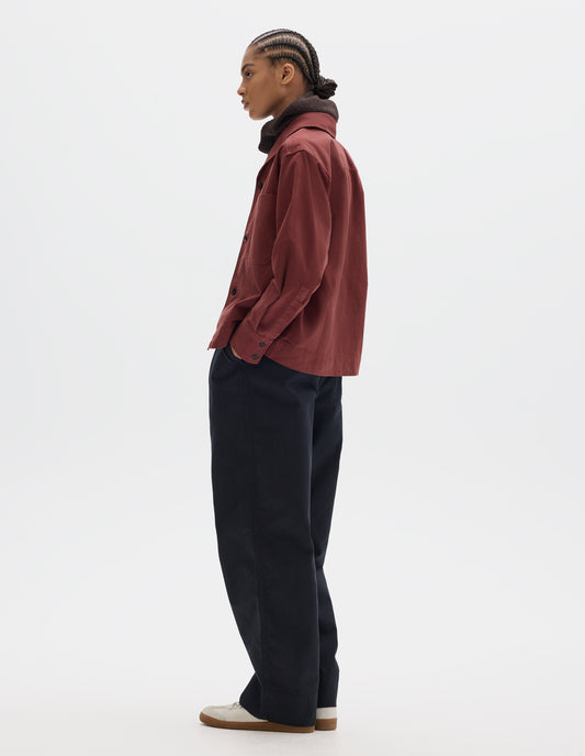 Full body image, front side view of model wearing a reddish cotton shawl collar overshirt with a front button placket, a forward-positioned shoulder seam and shirt cuffs with double button fastening. Styled with a brown wool hood, pleated trousers, off white suede trainers.