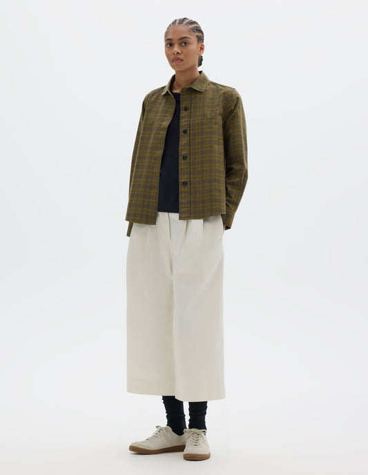 Full body image, front view of model wearing a green tonal check cotton shirt with a stitch-through raised placket and a curved hem. Styled open, over a black t shirt and worn with white pleated culotte trousers, black socks and white suede trainers.