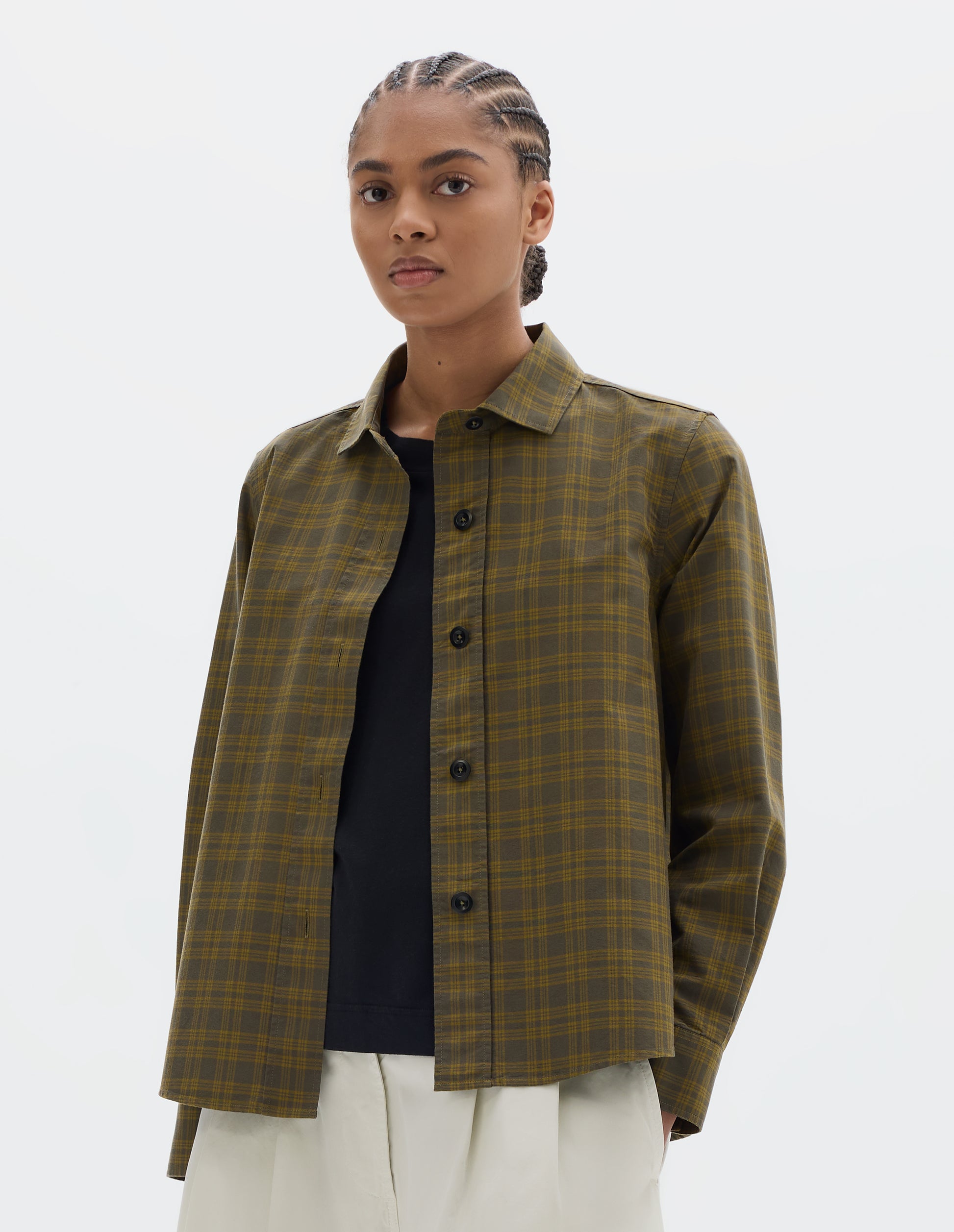 Cropped body image, front view of model wearing a green tonal check cotton shirt with a stitch-through raised placket and a curved hem. Styled open, over a black t shirt and worn with white pleated culotte trousers.