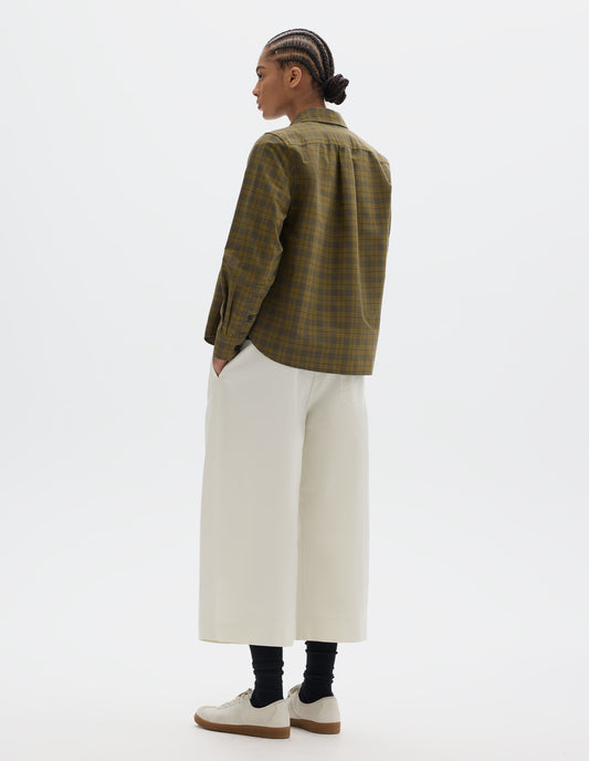 Full body image, back view of model wearing a green tonal check cotton shirt with a box pleat at the back yoke, button-through cuff gauntlet and a curved hem. Styled with white pleated culotte trousers, black socks and white suede trainers.