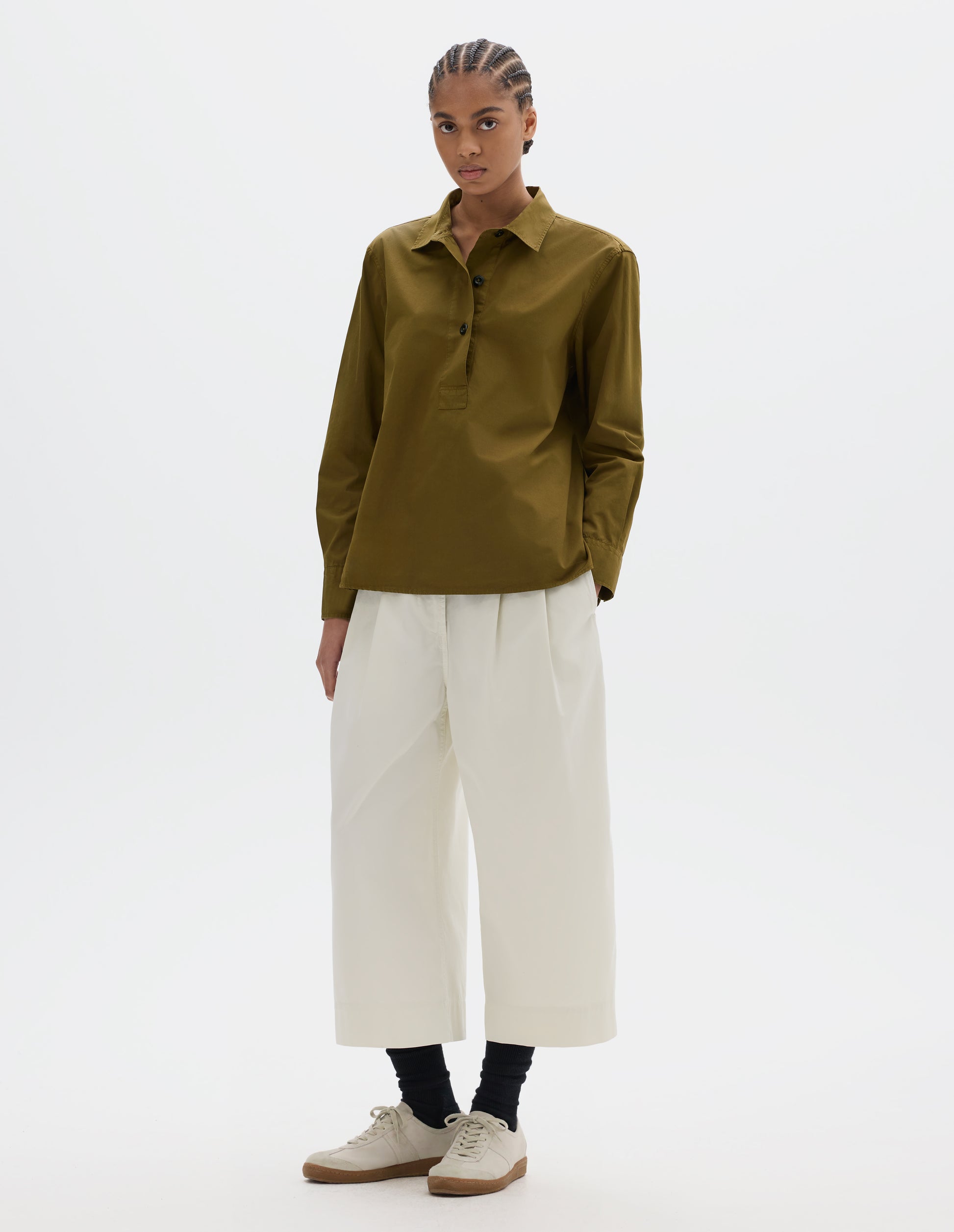Full body image, front view of model wearing an A-line brownish green cotton shirt, with dark buttons. Styled with white pleated cropped trousers, black socks and white suede trainers.
