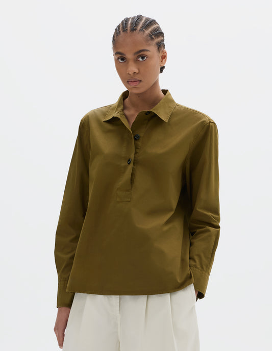 Cropped body image, front view of model wearing an A-line brownish green cotton shirt, with dark buttons. Styled with white pleated trousers.