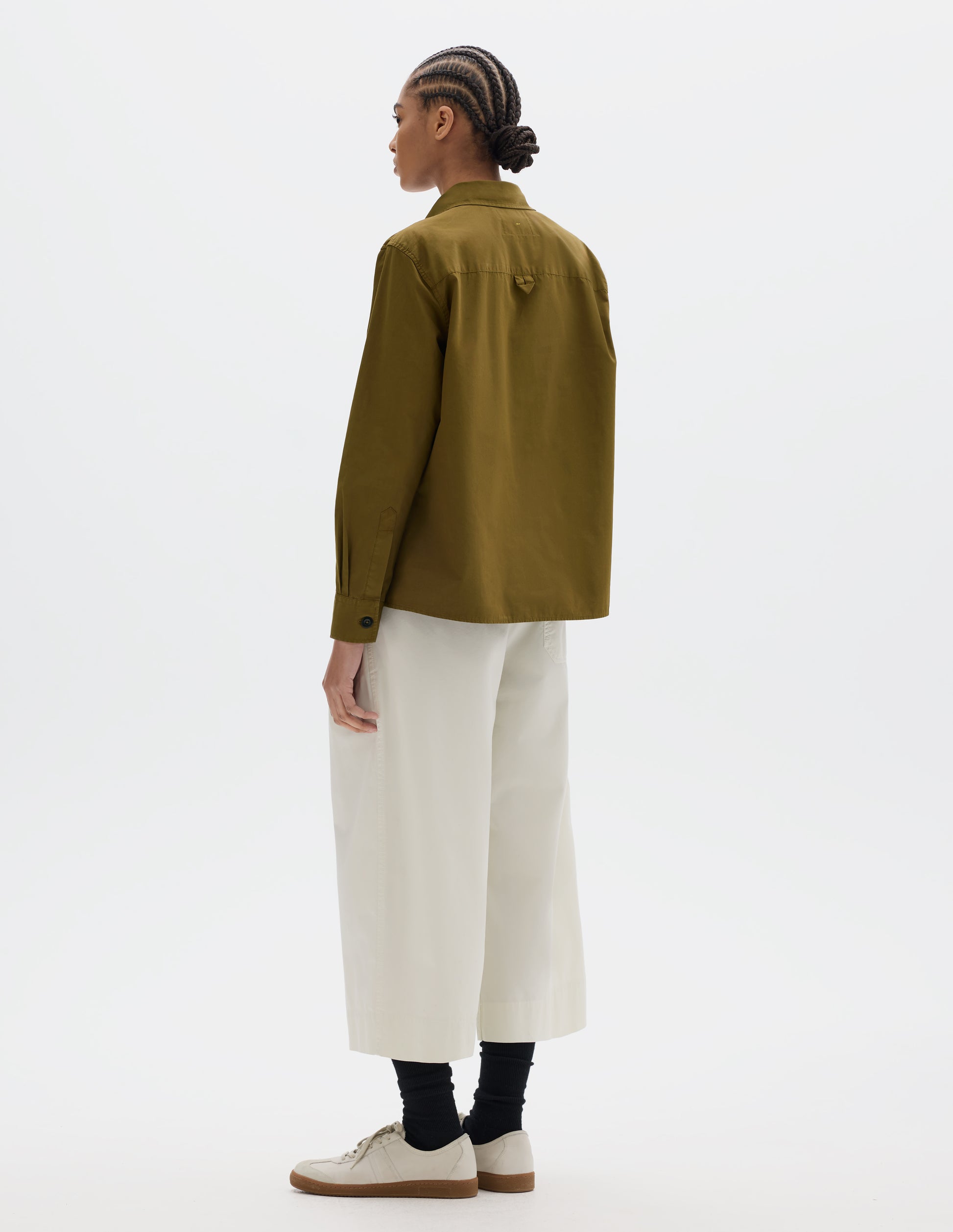 Full body image, back view of brownish green cotton shirt with hanger loop detail on back yoke. Styled with white pleated cropped trousers, black socks and white suede trainers.