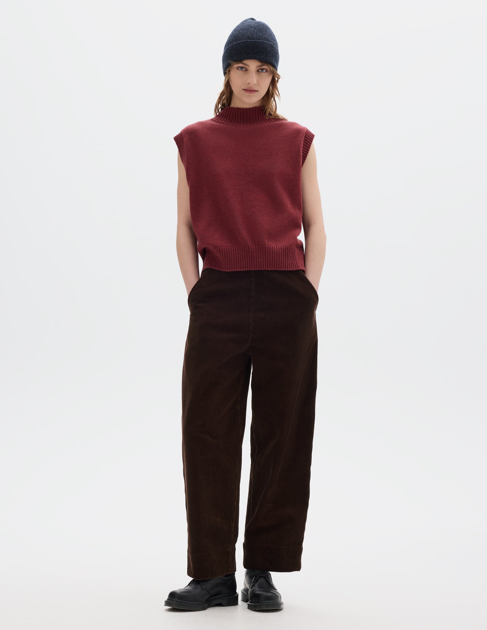 Full body image, front view of model wearing a reddish wool cotton slipover with a high rib neck, rib armholes and hem and split stepped hem. Styled with a dark grey wool beanie and dark brown corduroy trousers and black leather shoes.