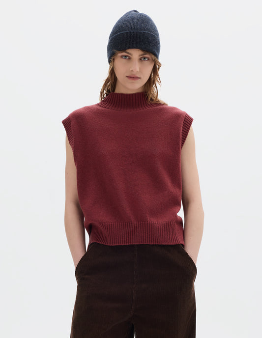 Cropped body image, front view of model wearing a reddish wool cotton slipover with a high rib neck, rib armholes and hem and split stepped hem. Styled with a dark grey wool beanie and dark brown corduroy trousers.