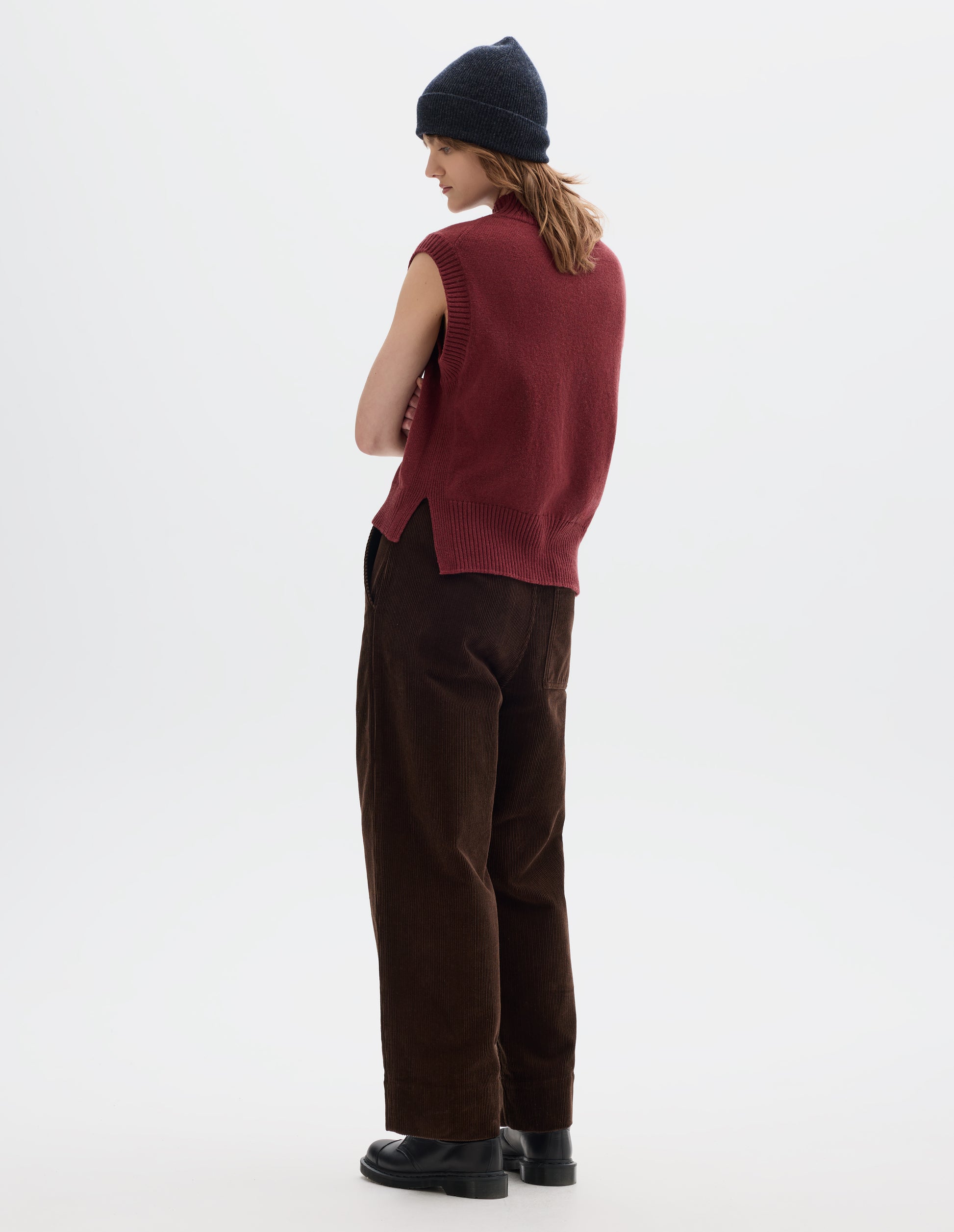 Full body image, back view of model wearing a reddish wool cotton slipover with a high rib neck, rib armholes and hem and split stepped hem. Styled with a dark grey wool beanie and dark brown corduroy trousers and black leather shoes.