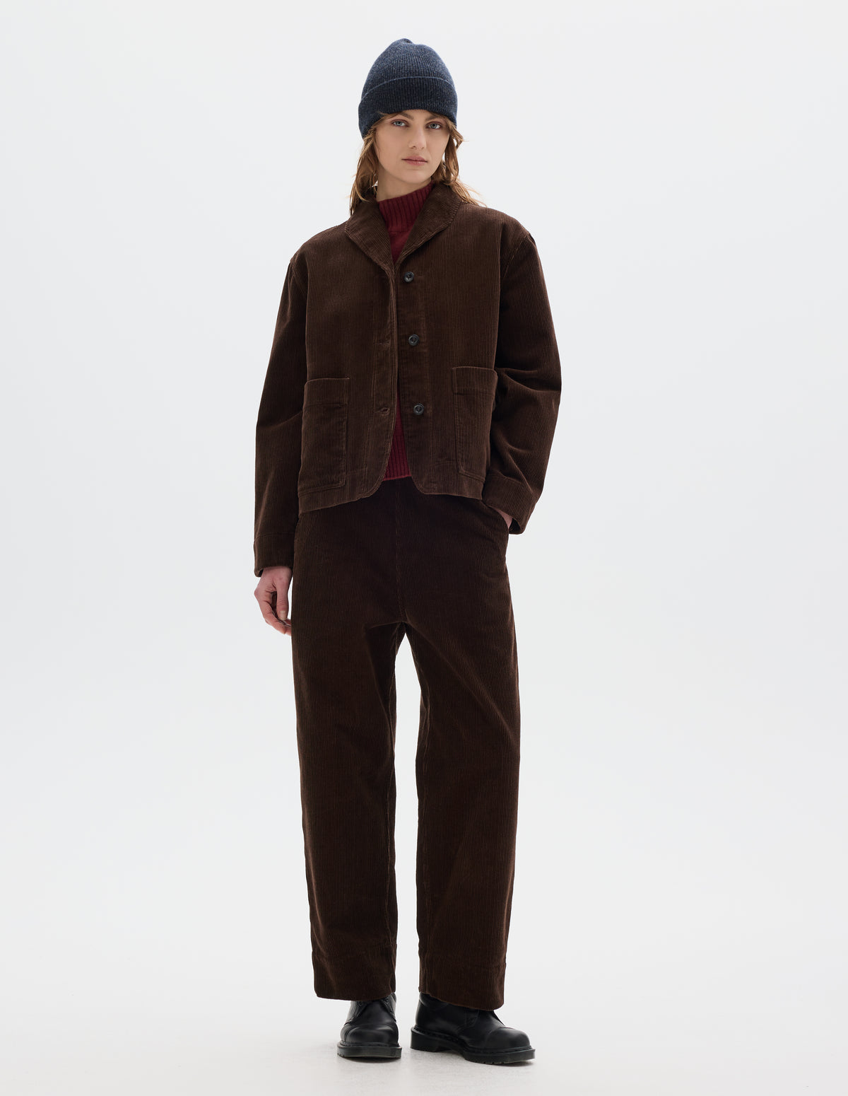 Full body image, front view of model wearing a brown corduroy jacket with two front patch pockets, stitch-through placket and a one piece collar. Styled with matching cord trousers, dark grey beanie, red wool high neck slipover and black leather shoes.