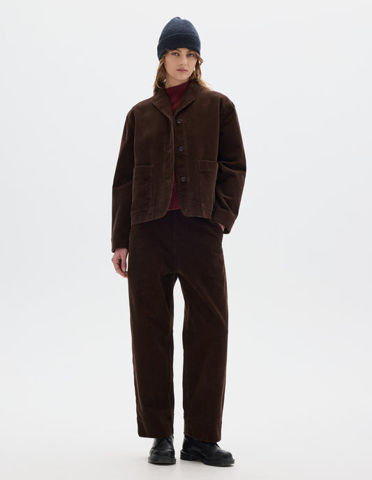 Full body image, front view of model wearing a brown corduroy jacket with two front patch pockets, stitch-through placket and a one piece collar. Styled with matching cord trousers, dark grey beanie, red wool high neck slipover and black leather shoes.