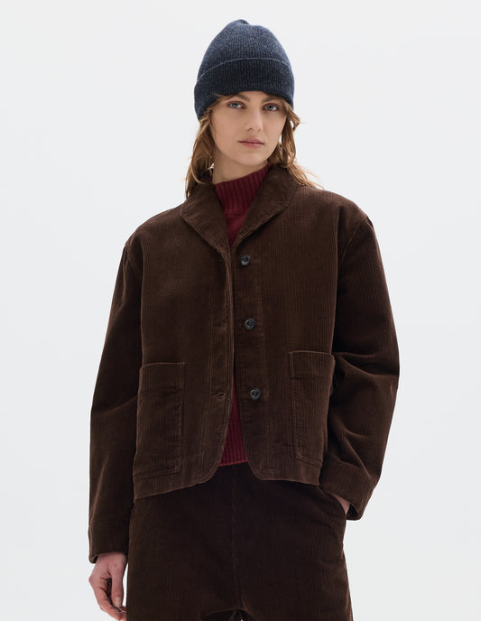 Cropped body image, front view of model wearing a brown corduroy jacket with two front patch pockets, stitch-through placket and a one piece collar. Styled with matching cord trousers, dark grey beanie and a red wool high neck slipover.