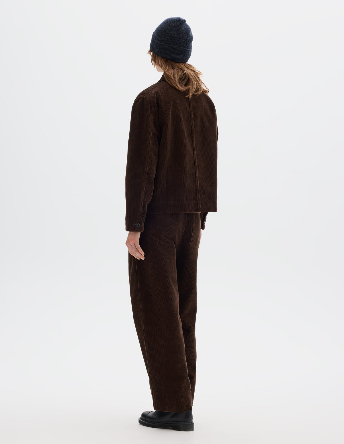 Full body image, back view of model wearing a brown corduroy jacket with shirt cuffs. Styled with matching cord trousers, dark grey beanie and black leather shoes.