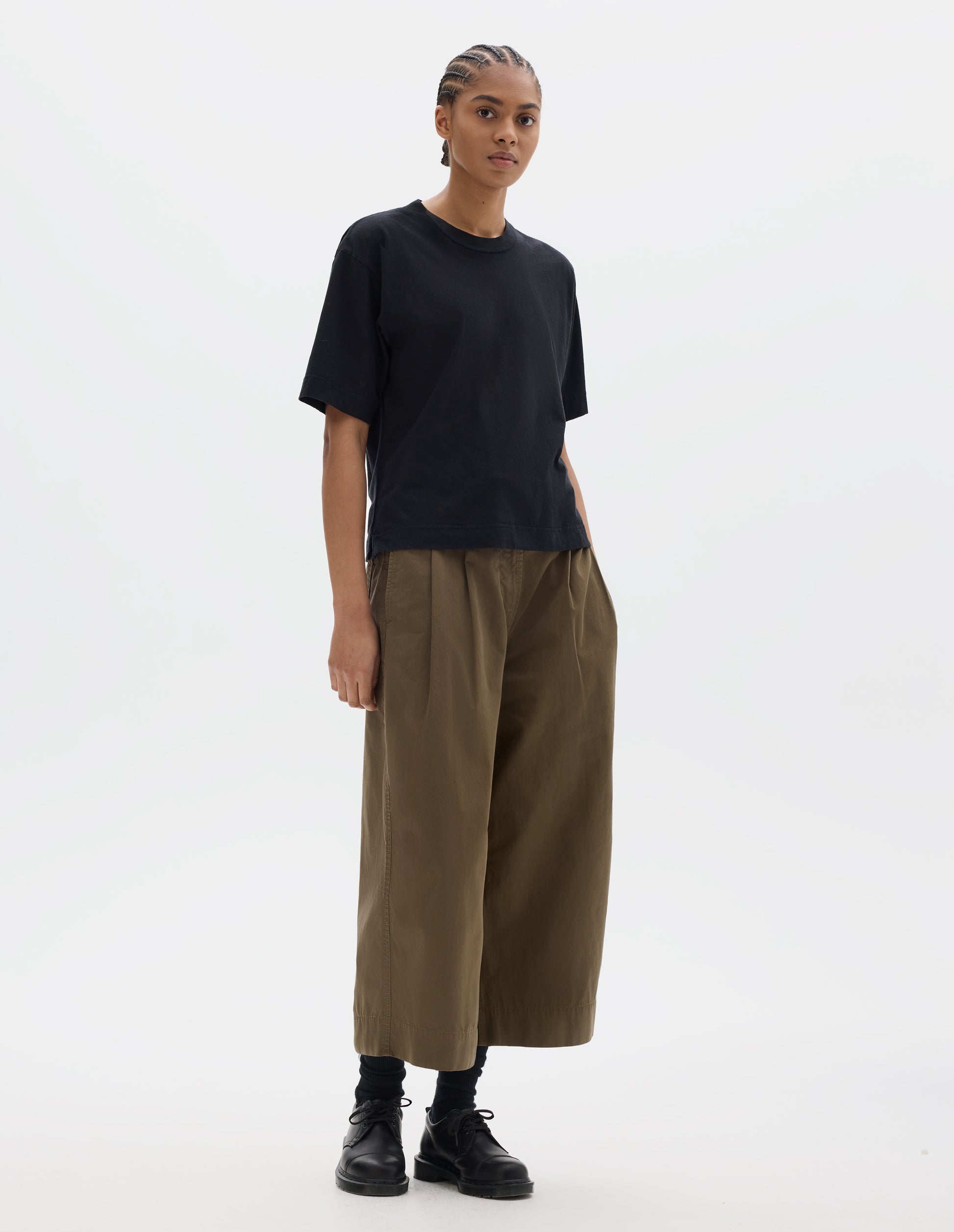 Full body image, front view of model wearing black short sleeve cotton t-shirt with contrast rib on neck. Styled with green brown pleated cropped trousers, black socks and leather shoes.