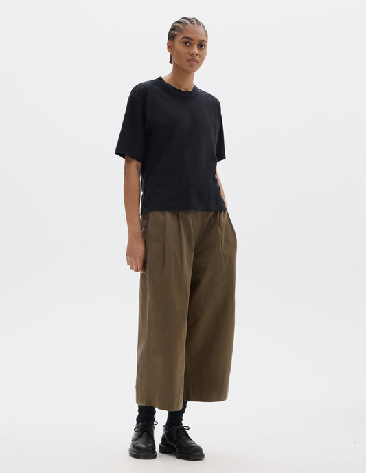 Full body image, front view of model wearing black short sleeve cotton t-shirt with contrast rib on neck. Styled with green brown pleated cropped trousers, black socks and leather shoes.