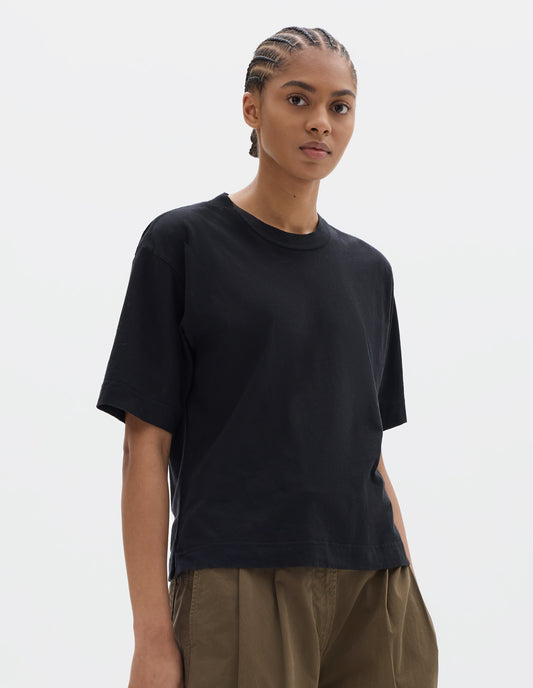 Cropped body image, front view of model wearing black short sleeve cotton linen t-shirt with contrast rib on neck. Styled with green brown pleated trousers.