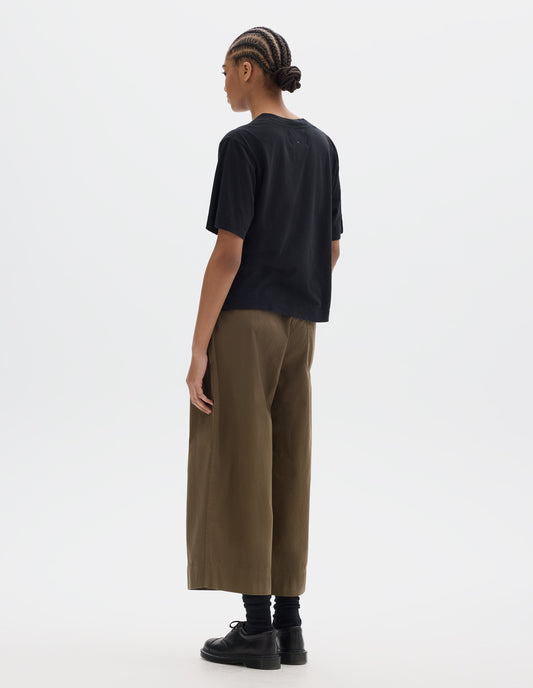 Full body image, back view of model wearing black short sleeve cotton t-shirt with contrast rib on neck and stitch detailing on back shoulder. Styled with green brown cropped trousers, black socks and leather shoes.