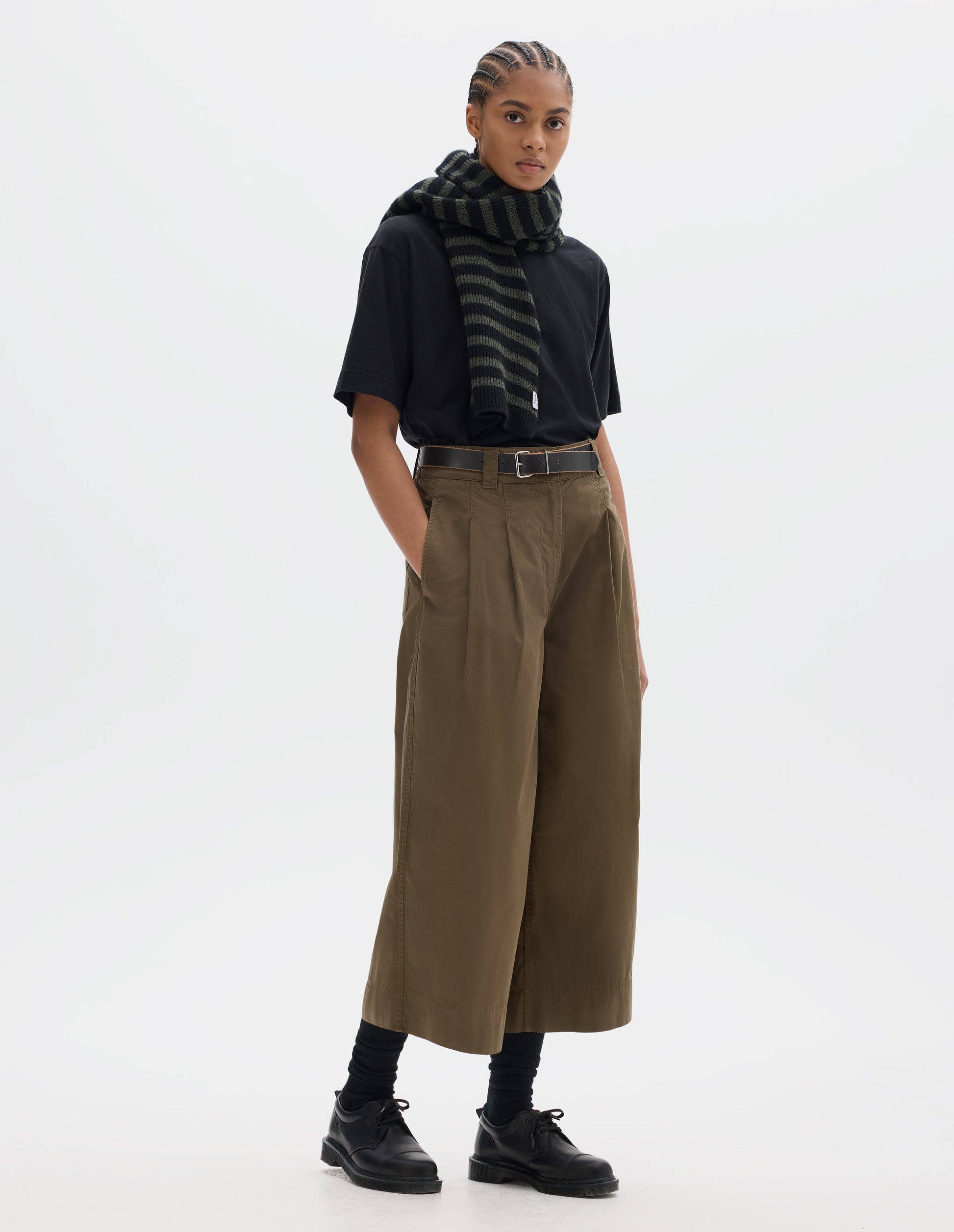 Full body image, front view of model wearing a black short sleeve t shirt styled with a green brown pleated flared cropped trousers. Worn with a striped wool scarf and black socks and black leather shoes.
