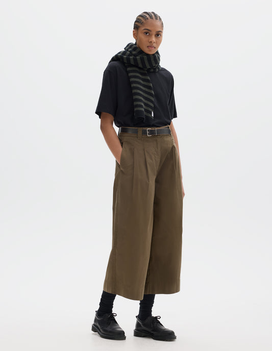 Full body image, front view of model wearing a black short sleeve t shirt styled with a green brown pleated flared cropped trousers. Worn with a striped wool scarf and black socks and black leather shoes.