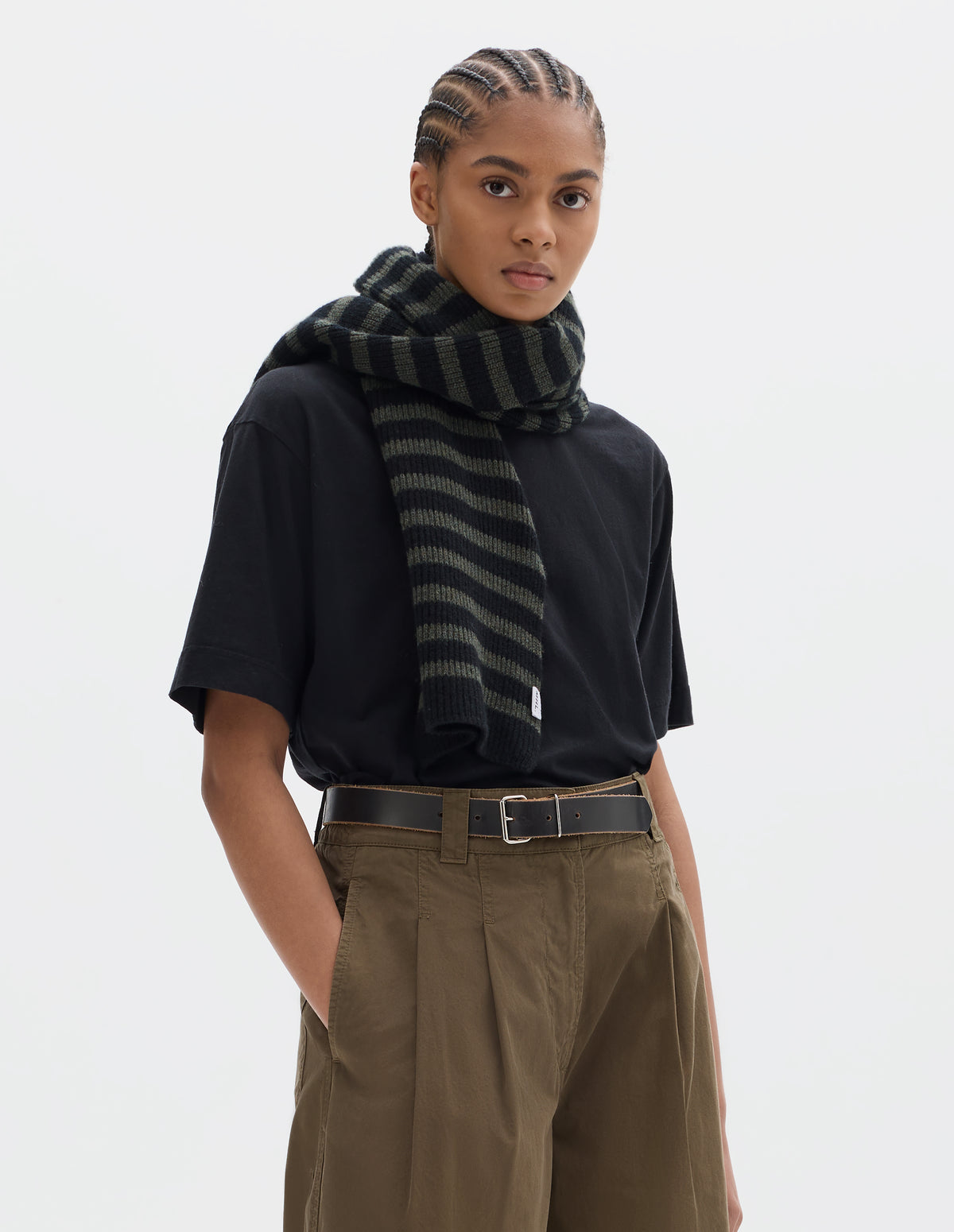 Cropped body image, front view of model wearing a striped black and brownish green wool scarf, a black short sleeve t shirt and a khaki green pleated trousers worn with a black leather belt.
