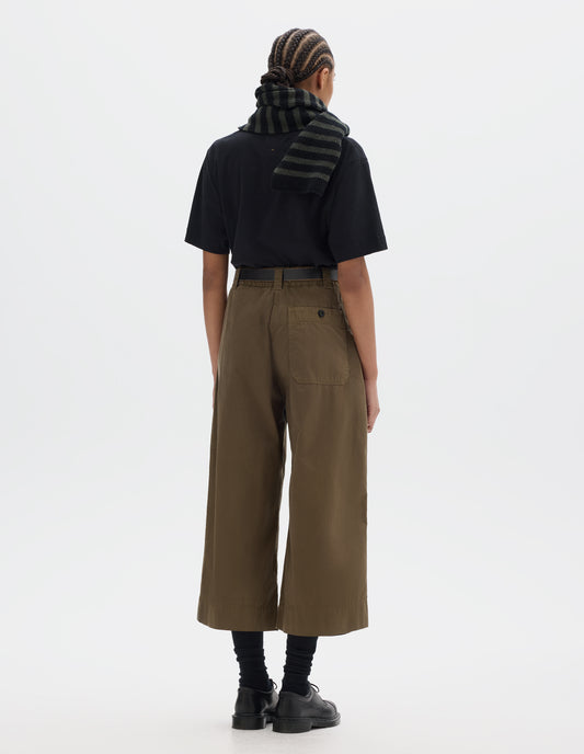 Full body image, back view of model wearing a black short sleeve t shirt, styled with a green brown flared cropped trousers. Worn with a striped wool scarf, black socks and black leather shoes.
