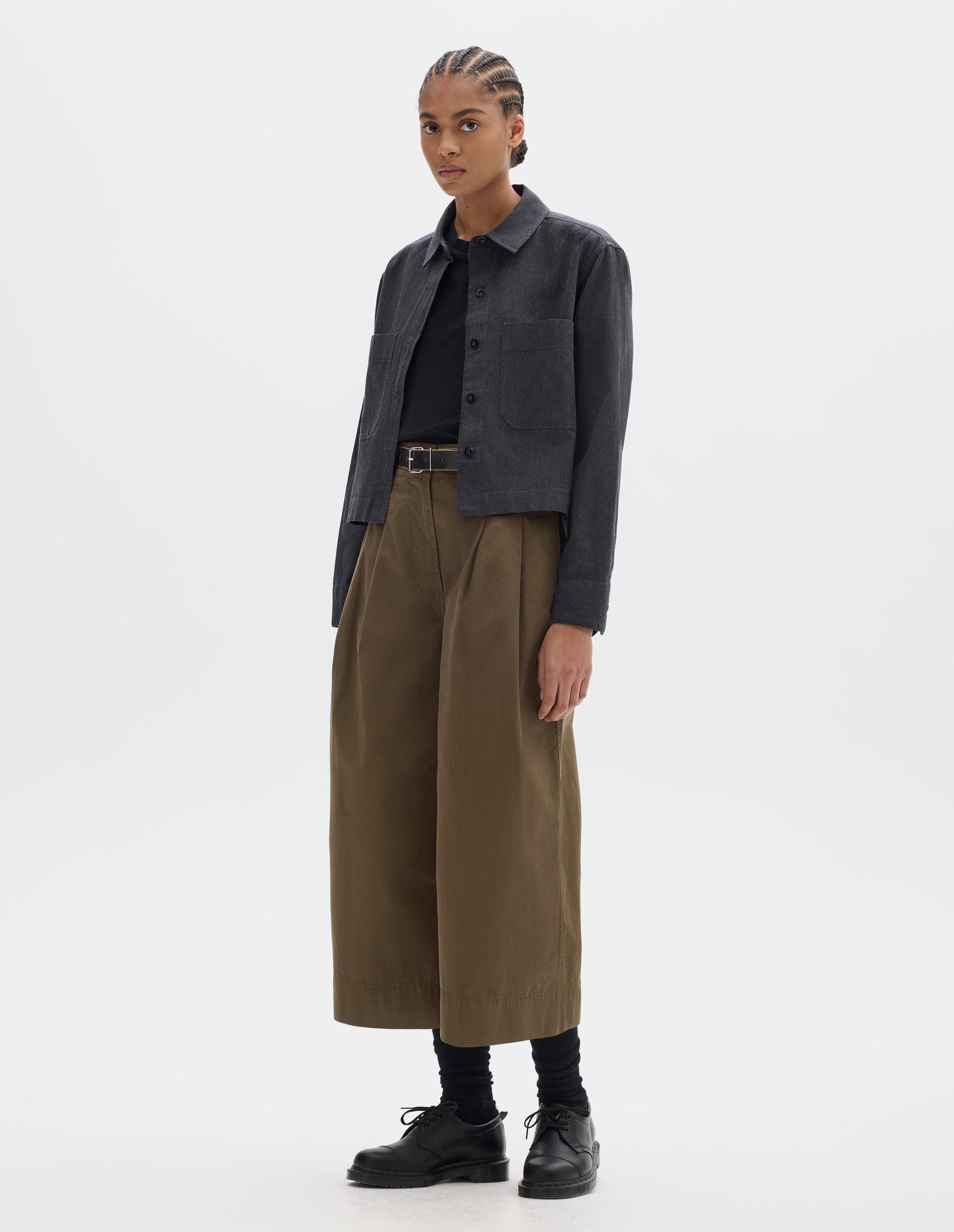 Full body image, front view of model wearing a dark grey cotton shirt, with patch chest pockets and dark buttons. Styled over a black t shirt, with greenish brown pleated culotte trousers and black leather belt, black socks and leather shoes.