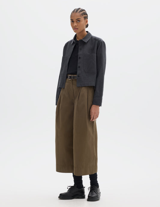 Full body image, front view of model wearing a dark grey cotton shirt, with patch chest pockets and dark buttons. Styled over a black t shirt, with greenish brown pleated culotte trousers and black leather belt, black socks and leather shoes.