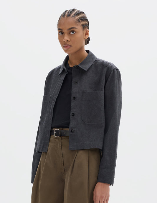 Cropped body image, front view of model wearing a dark grey cotton shirt, with patch chest pockets and dark buttons. Styled over a black t shirt, with greenish brown pleated culotte trousers and black belt.
