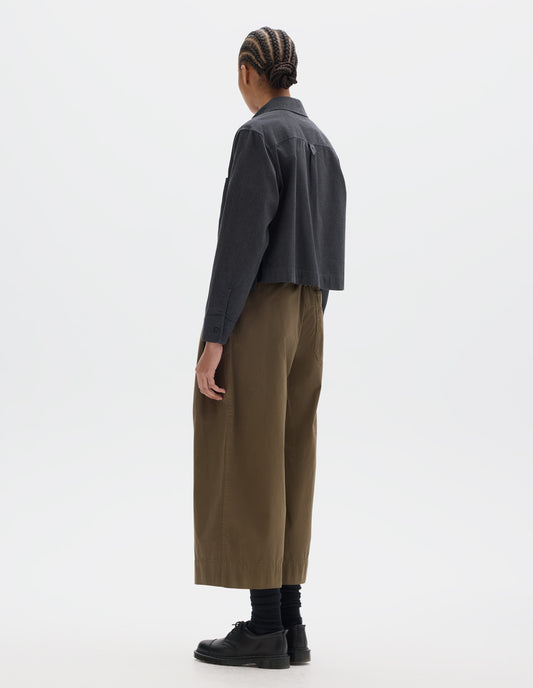 Full body image, back view of model wearing a dark grey cotton shirt, grown-on cuffs, and a back yoke with a box pleat and external hem loop detailing. Styled with greenish brown culotte trousers and black socks leather shoes.