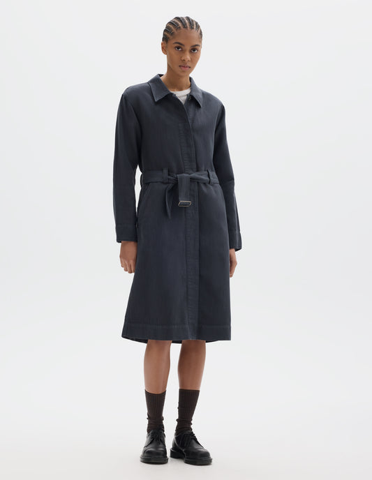 Full body image, front view of model wearing a dark navy cotton linen overall dress with a fly front with button fastening, slanted pockets and a removable belt with a metal slide adjuster. Styled over a white t shirt and worn with black socks and black leather shoes.