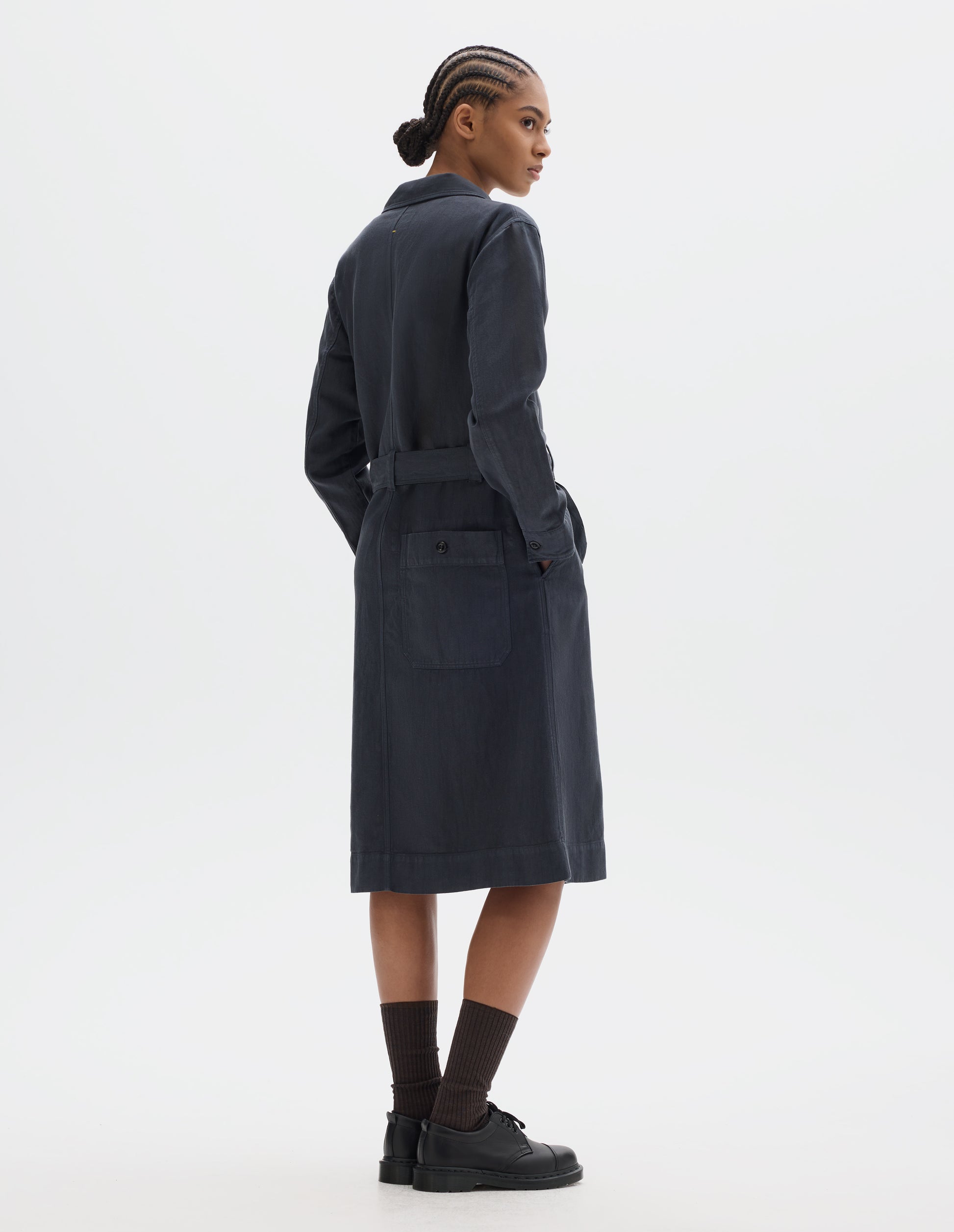 Full body image, back view of model wearing a dark navy cotton linen overall dress with slanted pockets, button-fastening split cuffs, button-through patch pocket at the back and a removable belt. Styled with black socks and leather shoes.