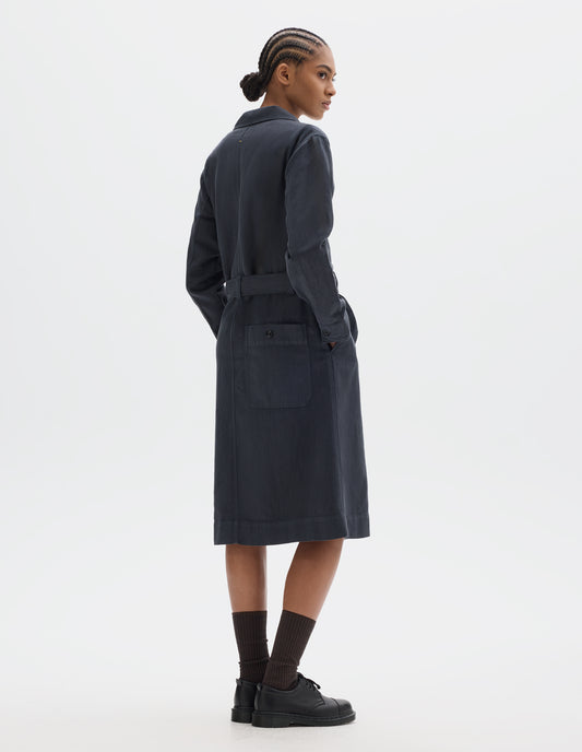 Full body image, back view of model wearing a dark navy cotton linen overall dress with slanted pockets, button-fastening split cuffs, button-through patch pocket at the back and a removable belt. Styled with black socks and leather shoes.