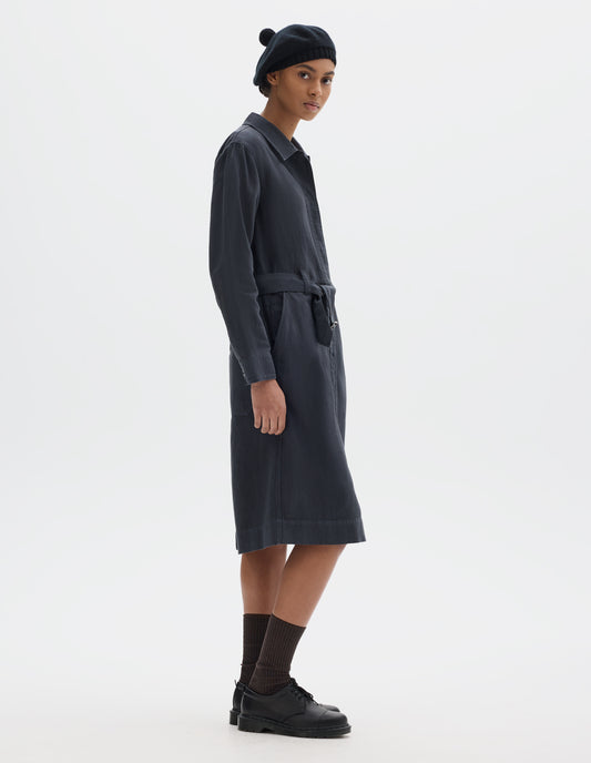 Full body image, front side view of model wearing a dark navy cotton linen overall dress with a fly front with button fastening, slanted pockets and a removable belt with a metal slide adjuster. Styled with a black bobble beret, black socks and leather shoes.