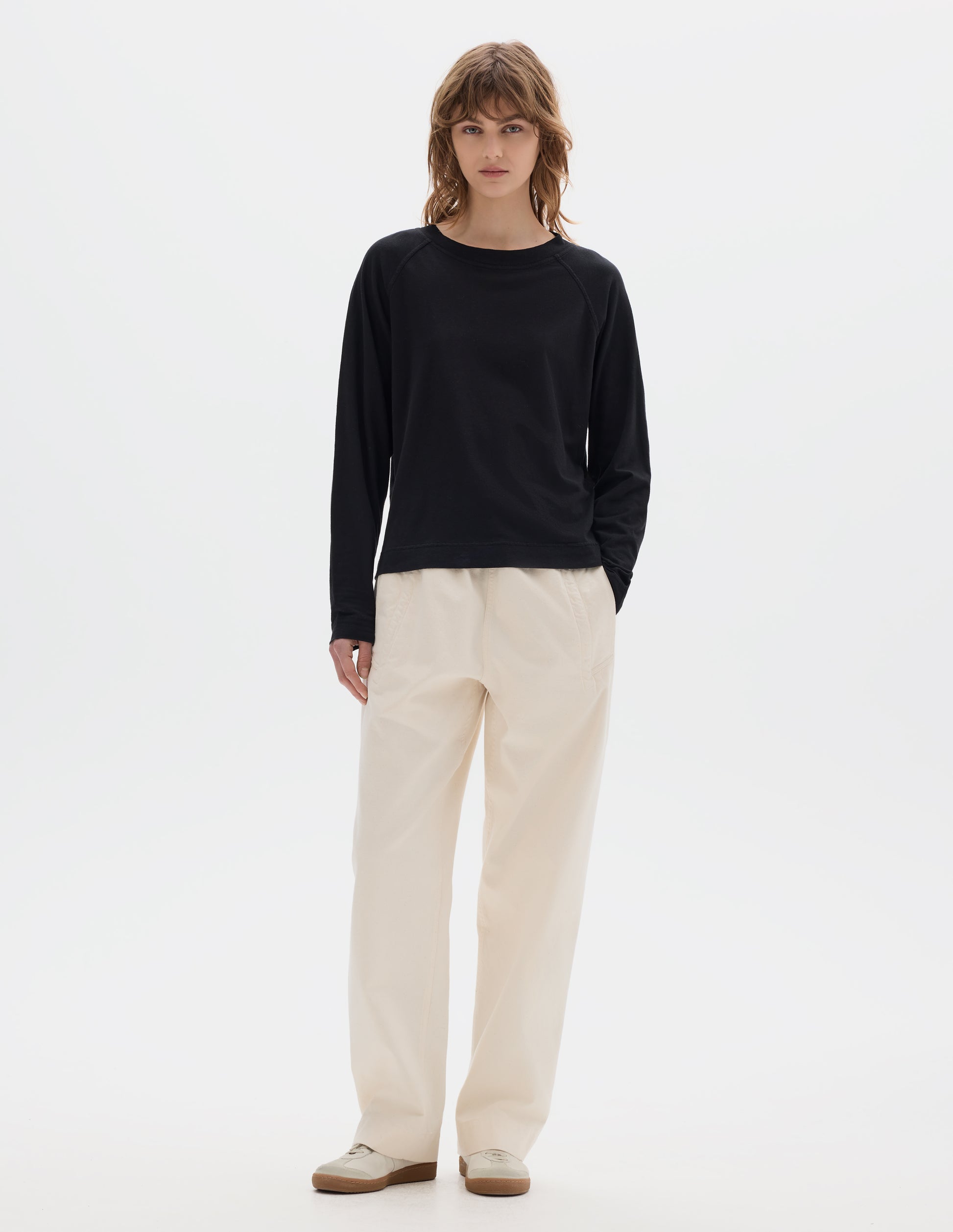 Full body image, front view of model wearing a black cotton linen raglan t shirt with a rib crew neck. Styled with jogger style off white trousers and suede trainers.