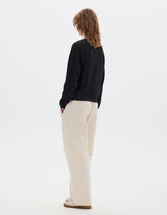 Full body image, back view of model wearing a black cotton linen raglan t shirt with a rib crew neck. Styled with jogger style off white trousers and suede trainers.