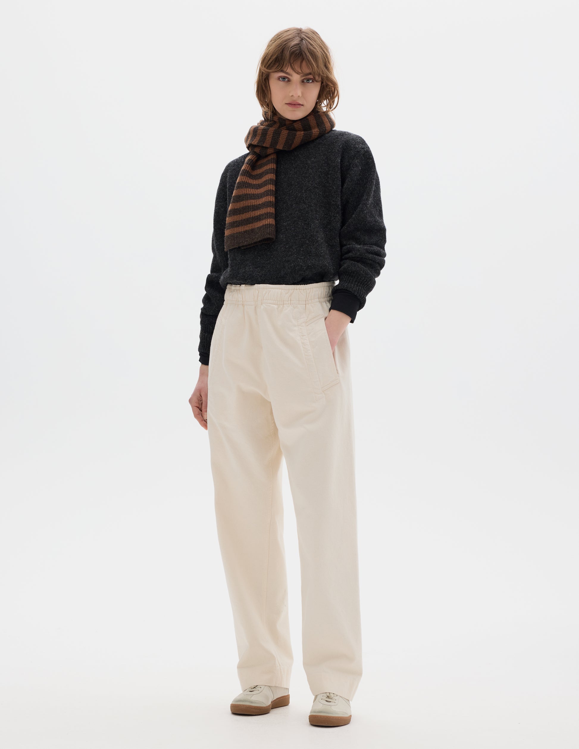 Full body image, front view of model wearing off white joggers with elasticated waist and deep welt pockets. Styled with a dark grey wool sweater, tonal brown stripe scarf and off white suede trainers.