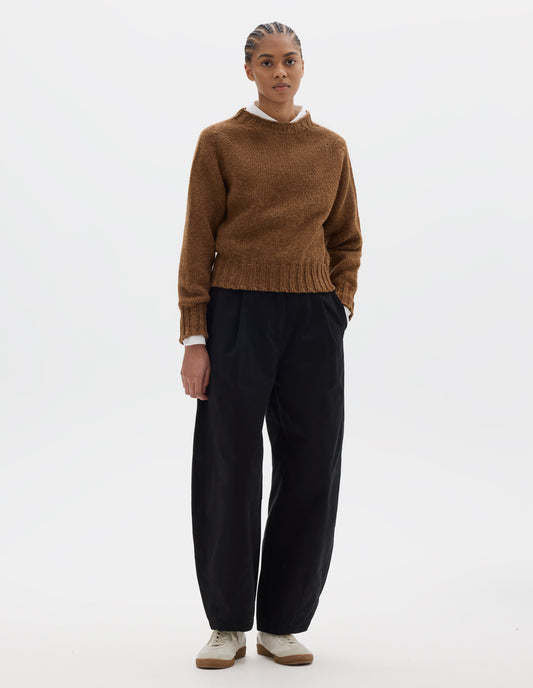 Full body image, front view of model wearing a orangey brown crew neck wool jumper with saddle sleeves and contrasting rib on the neck, cuffs and hem. Styled over a white shirt, black pleated barrel leg trousers and off white suede trainers.