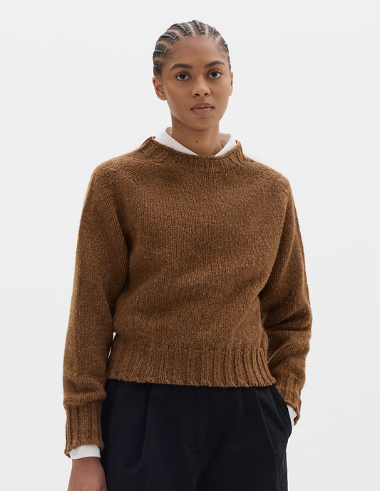 Cropped body image, front view of model wearing a orangey brown crew neck wool jumper with saddle sleeves and contrasting rib on the neck, cuffs and hem. Styled over a white shirt and black pleated trousers.