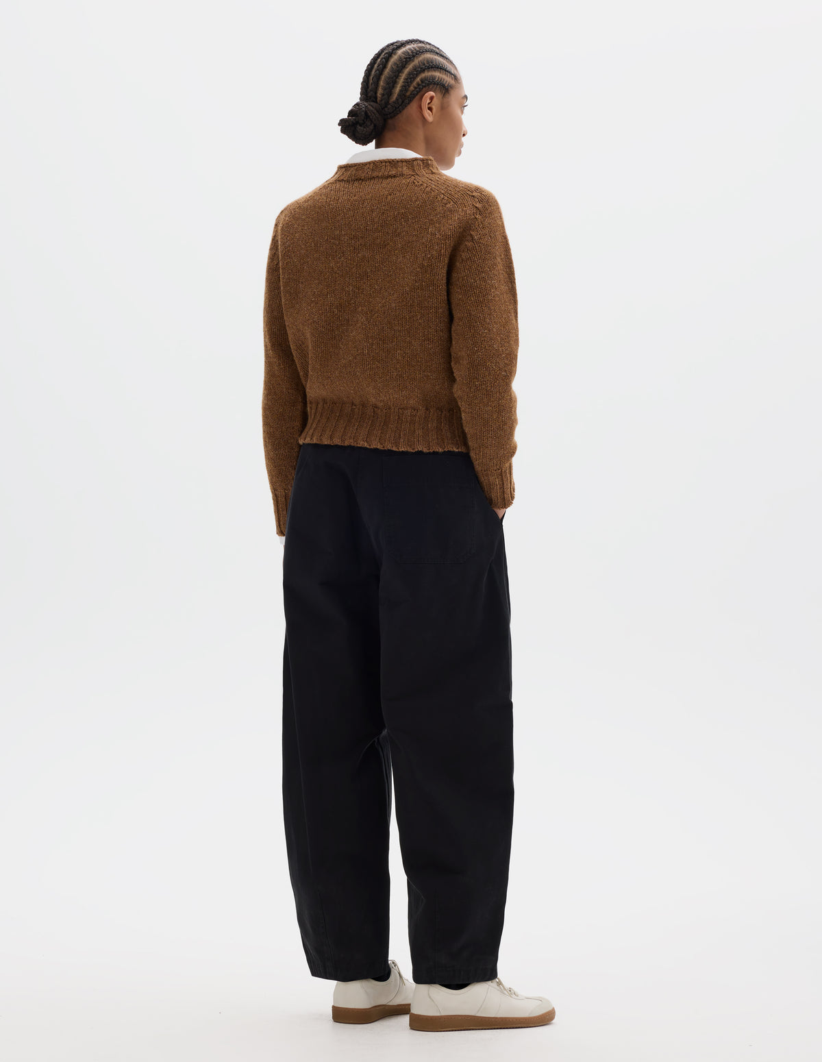 Full body image, back view of model wearing a orangey brown crew neck wool jumper with saddle sleeves and contrasting rib on the neck, cuffs and hem. Styled over a white shirt, black barrel leg trousers and off white suede trainers.