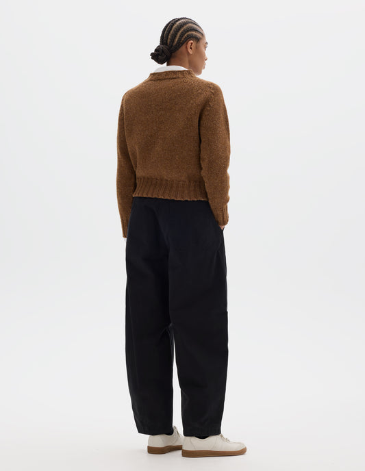 Full body image, back view of model wearing a orangey brown crew neck wool jumper with saddle sleeves and contrasting rib on the neck, cuffs and hem. Styled over a white shirt, black barrel leg trousers and off white suede trainers.