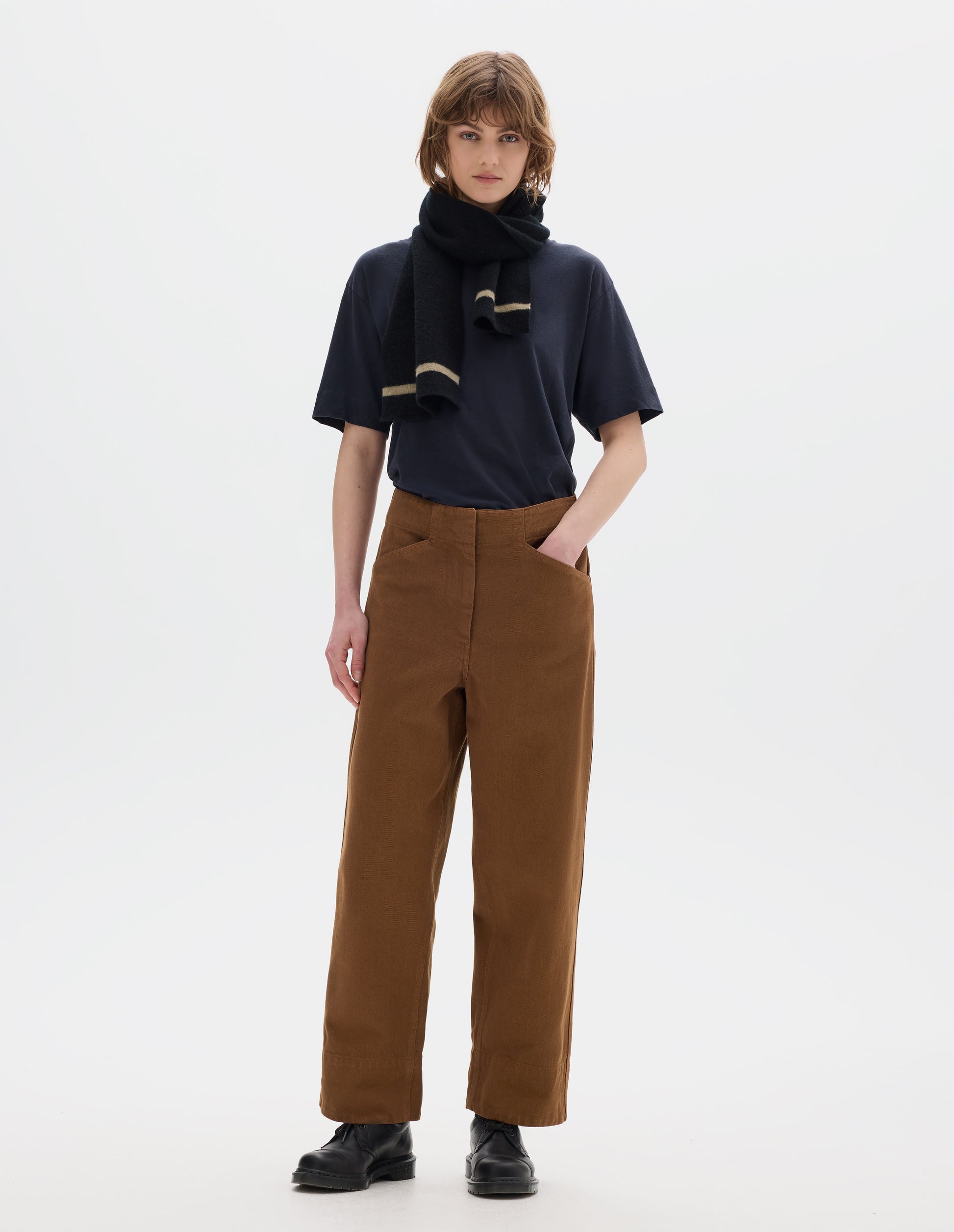 Full body image, front view of model wearing a dark short sleeve t shirt, brown cotton trousers, with front pockets and deep hems. Styled with a black wool scarf with beige edge stripe and black leather shoes.